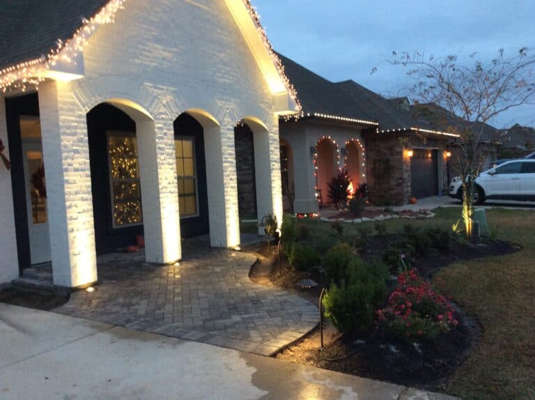 A suburban house in Baton Rouge with a white brick exterior is beautifully decorated with outdoor lighting for the holidays. The front porch has illuminated archways, and the yard features well-maintained landscaping with shrubs and flowers. Another house and a white car are visible.