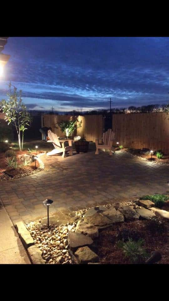 A tranquil backyard in Baton Rouge at dusk with two wooden chairs facing each other on a stone patio. Warm outdoor lighting illuminates the space, highlighting plants, a small garden, and a wooden fence. The sky is a deep blue with scattered clouds.