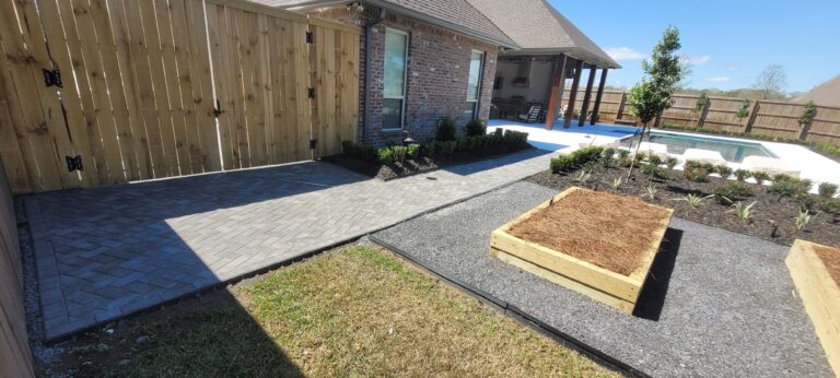 A backyard with a brick house, wooden fence, paved walkway, garden beds, and a swimming pool. Neat landscaping includes shrubs and a small tree. The garden beds are raised and filled with soil, perfect for an herb garden. A covered patio area is visible in the background.