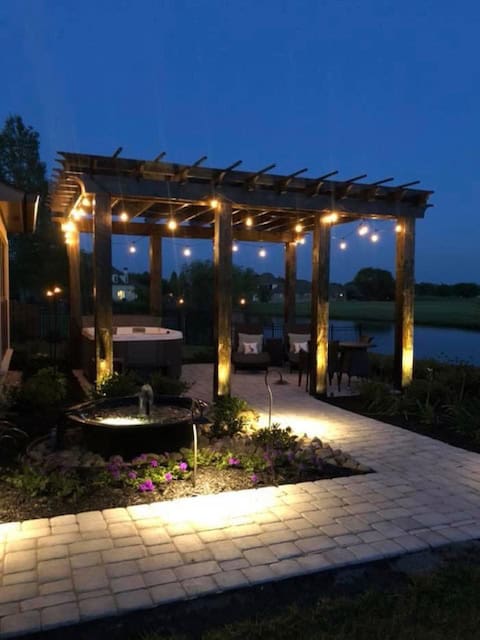 Outdoor patio area at dusk in Baton Rouge, featuring a wooden pergola with string lights, a hot tub, and cozy seating. The space is enhanced by outdoor lighting and ground lights, highlighting a small fountain and vibrant flowerbed along a paved walkway.