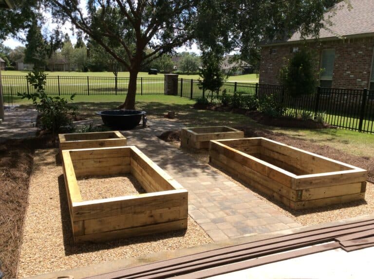 A backyard vegetable garden with four large wooden raised beds is arranged on and around a stone-paved pathway. There are trees and a grassy lawn in the background, with a brick house and black fence surrounding the area. Pine straw mulch covers the ground around this well-planned garden installation.