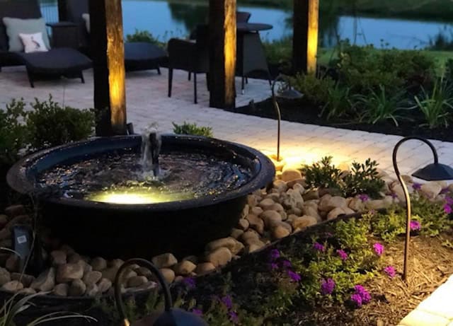 A cozy outdoor patio scene in Baton Rouge features a water fountain surrounded by rocks and enhanced with soft, warm outdoor lighting. The area is beautifully landscaped with greenery and purple flowers, overlooking a calm body of water in the background.