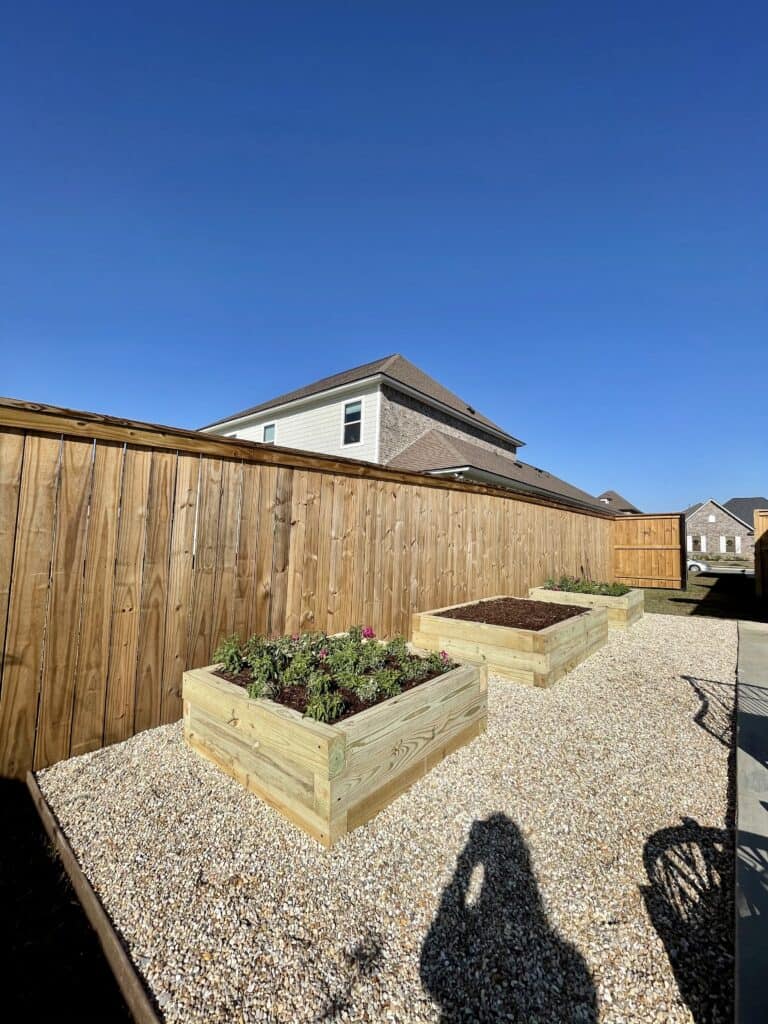 A backyard garden installation featuring two raised wooden beds filled with soil and plants. The vegetable garden is enclosed by a tall wooden fence, and gravel covers the ground. A shadow of a person taking the picture is visible on the gravel. A house is seen in the background under a clear blue sky.