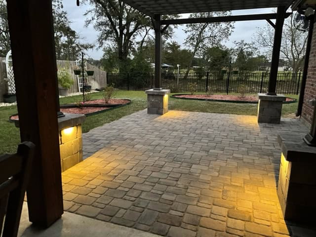 A backyard patio with stone pavers, illuminated by soft yellow outdoor lighting. The area is covered by a wooden pergola, with landscaped circular flower beds in the background and a black metal fence along the edge of the yard. Tall trees are visible beyond the fence, reminiscent of Baton Rouge’s serene landscapes.
