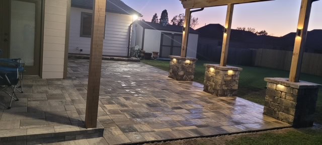 A backyard patio at dusk with stone pavers and square columns supporting a partially covered wooden pergola. The outdoor lighting illuminates the columns and casts a warm glow over the Baton Rouge evening. The yard beyond is grassy, with a wooden fence and silhouetted buildings.