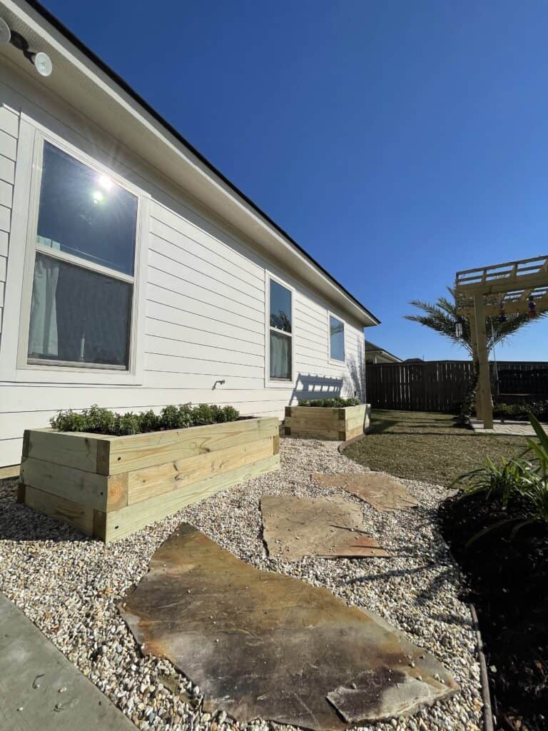A backyard with a white house showing two large windows. It features two wooden raised garden beds filled with plants, perfect for a vegetable garden. Stone pavers create a pathway on gravel, leading to a pergola in a grassy area. A clear blue sky is visible above.