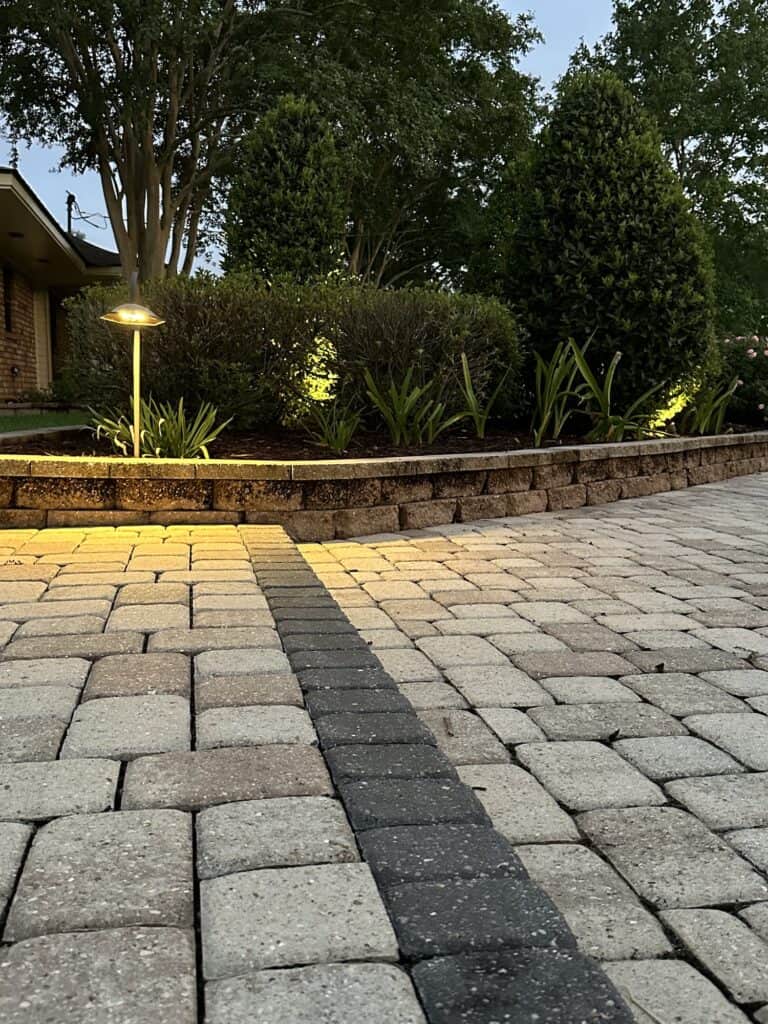 A paved patio with a dark stone stripe, trimmed shrubs, and Baton Rouge outdoor lighting illuminating the garden bed at dusk.