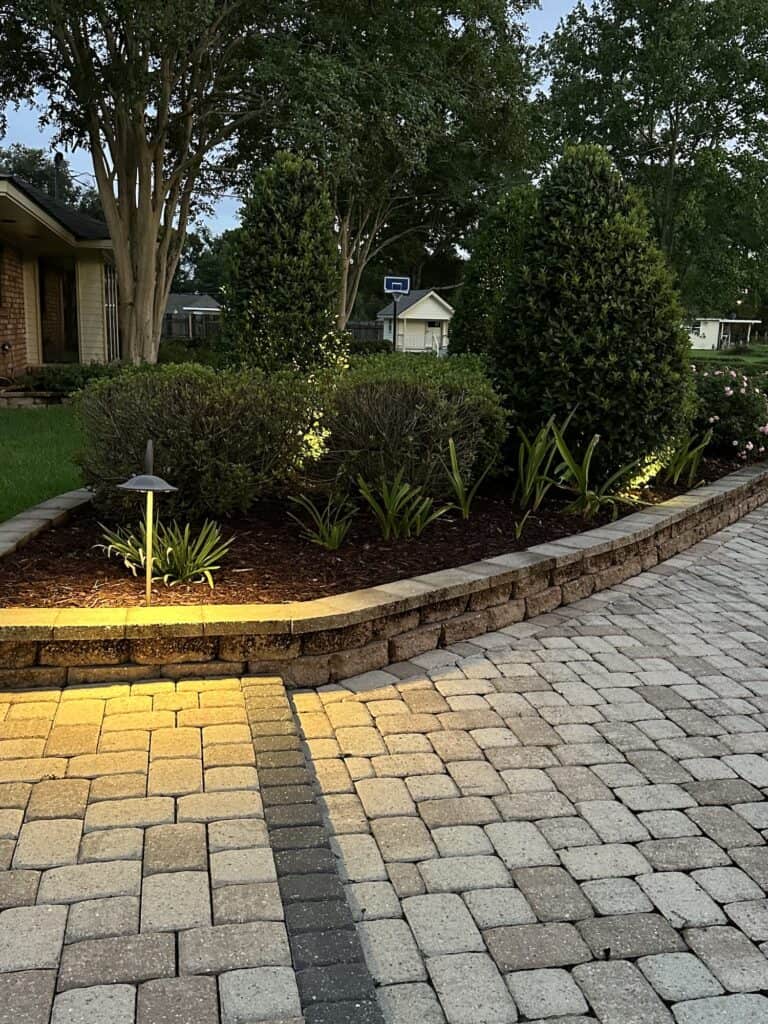 A landscaped garden bed with shrubs and small plants in Baton Rouge, bordered by a stone wall and enhanced by outdoor lighting at dusk.