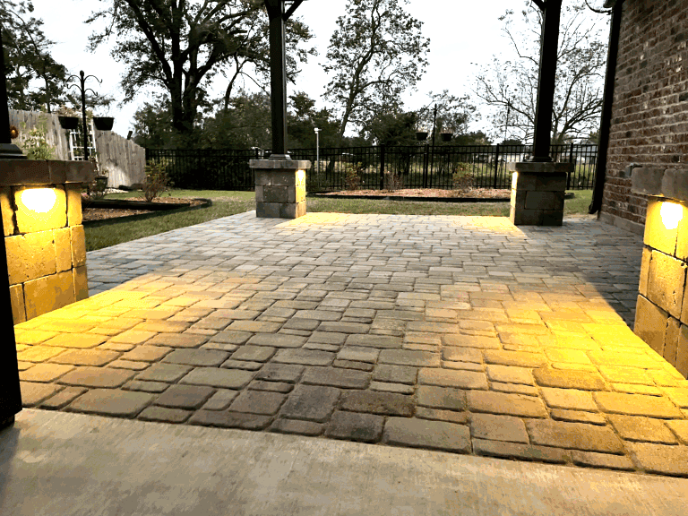 Brick patio with built-in outdoor lighting at dusk, bordered by grass and trees, set in scenic Baton Rouge with a black metal fence.