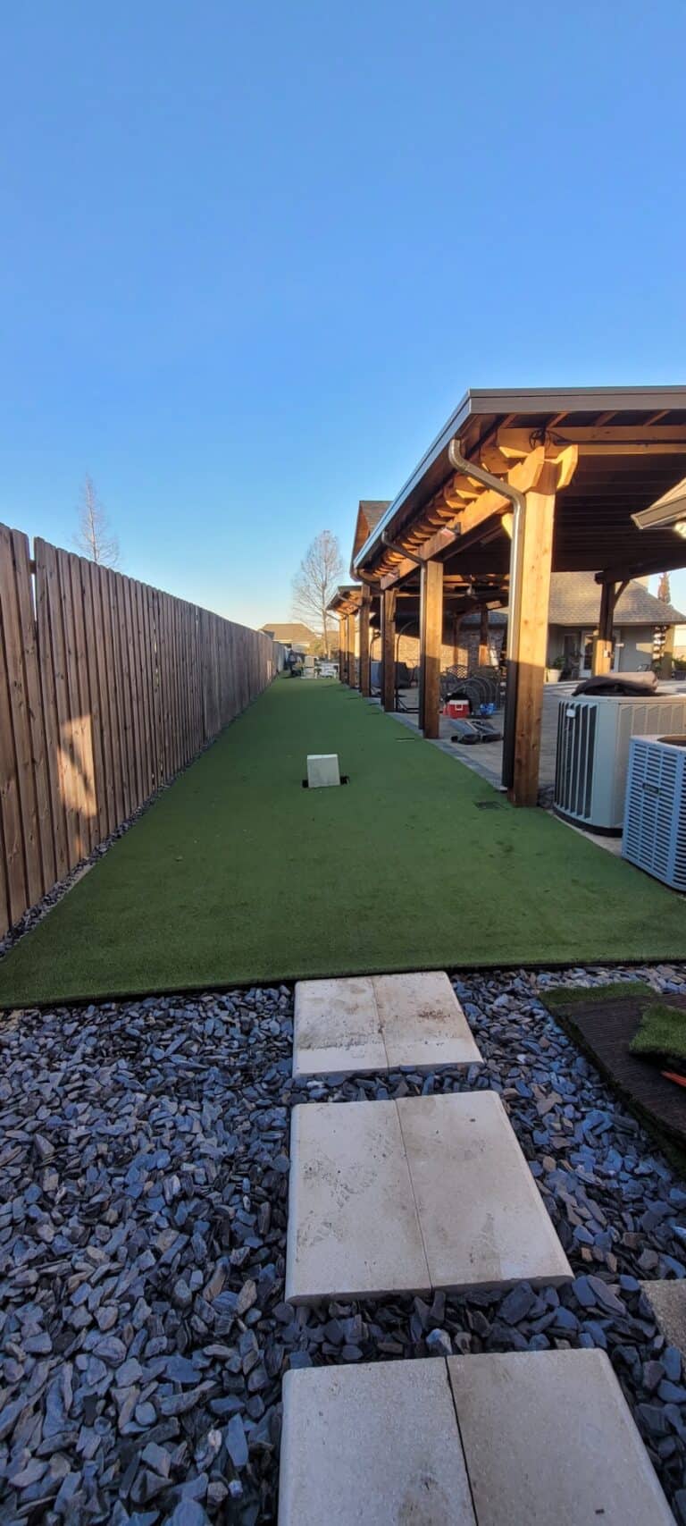 A backyard with artificial grass bordered by a wooden fence on the left and a covered patio on the right, under a clear sky.