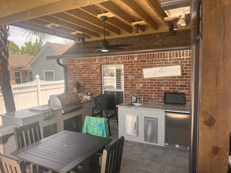 Covered outdoor kitchen with grill, sink, fridge, and dining table under a wooden pergola with stylish roof extensions and brick wall background.