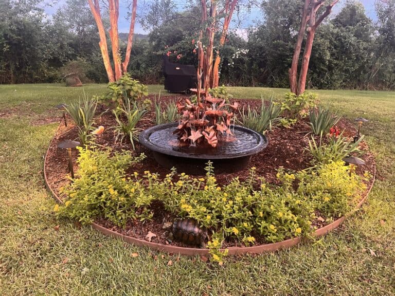 A circular garden bed with a metal water feature installation at its center, surrounded by flowers, plants, mulch, and bordered by grass.