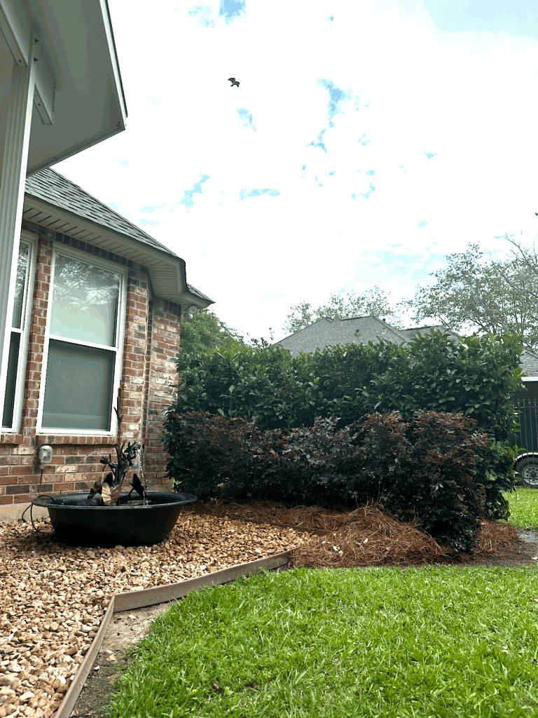 A brick house with a window, garden bed, bird bath water feature, and green grass under a partly cloudy sky.