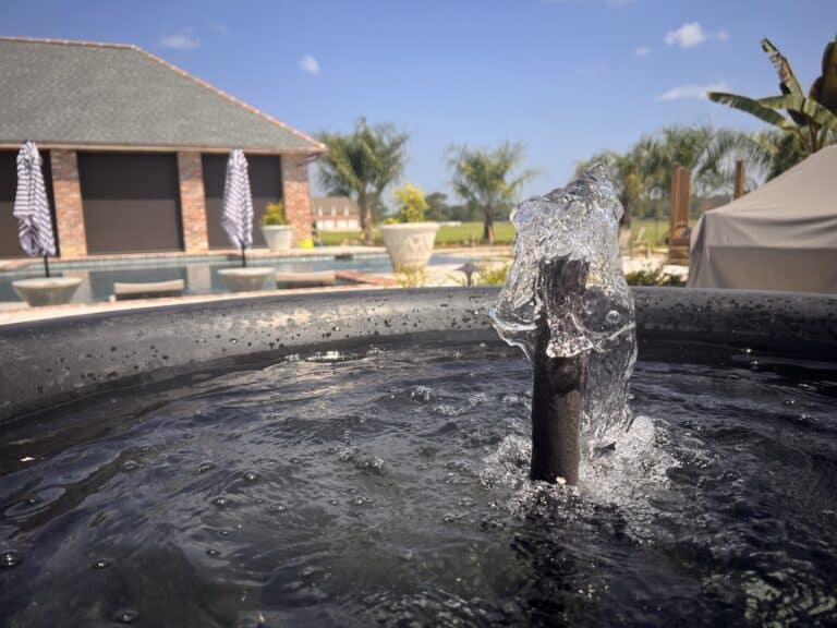 Close-up of a water feature installation in a black basin, with a pool, palm trees, and brick building in the background.