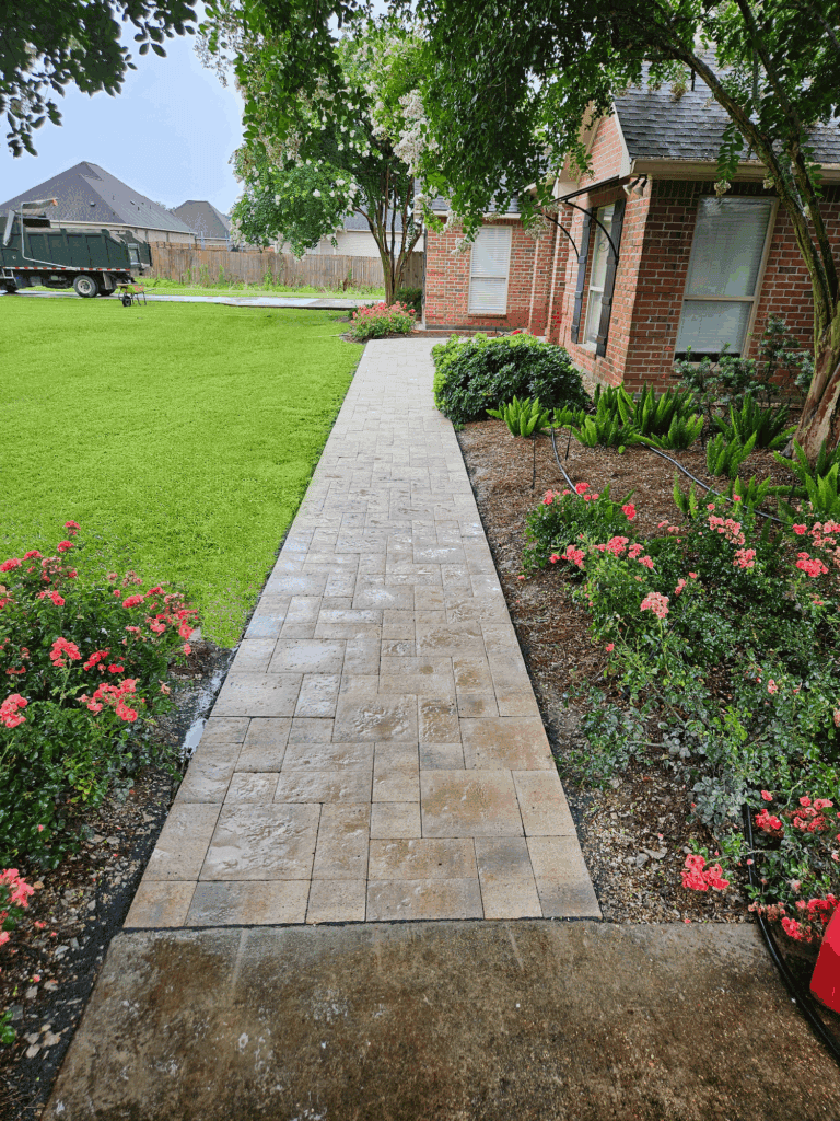 A paved walkway of decorative pavers leads to a brick house, bordered by a green lawn and vibrant pink and red flower beds.