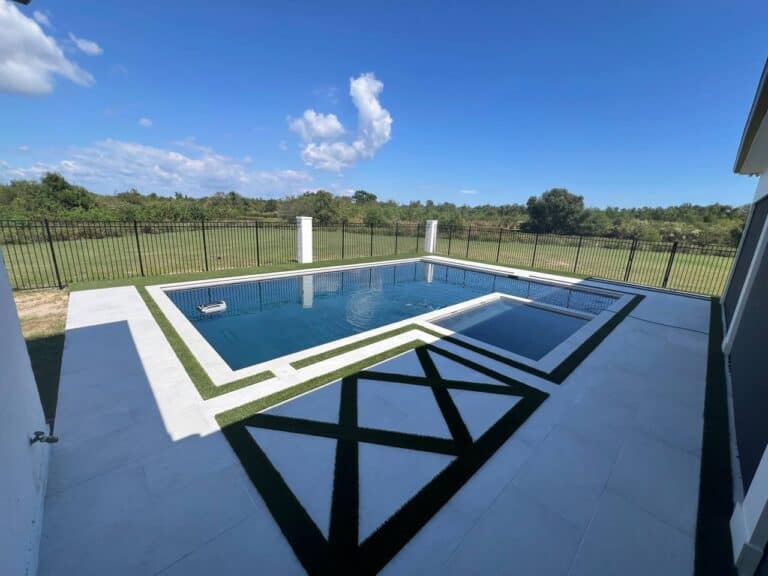 Rectangular backyard pool with hot tub, geometric tile, and black fence, featuring Synthetic Turf Installation beside a grassy field and blue sky.