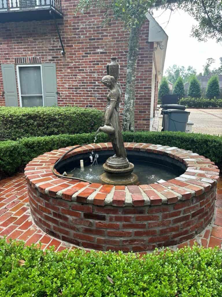 A brick circular water feature installation with a central statue, surrounded by green hedges in a residential courtyard.