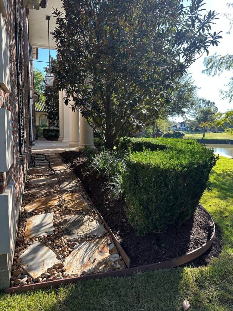 A natural stone pathway runs alongside a house, bordered by trimmed bushes and mulch beds, with a lake visible in the background.