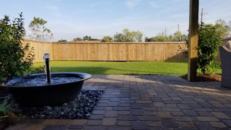 A backyard patio with a stone-tile floor features a small black fountain installed over rocks in the foreground, creating an elegant water feature. The yard boasts a well-maintained green lawn bordered by a tall wooden fence, with trees and power lines visible under the clear sky.
