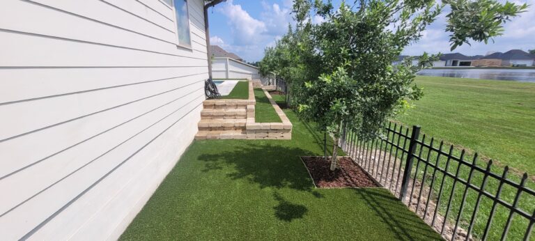A side yard with synthetic turf, a young tree planted in a small mulch bed, and a white house with a wooden fence. Stone steps and a low stone retaining wall lead to a raised area. In the background, there are more houses and a large grassy field.