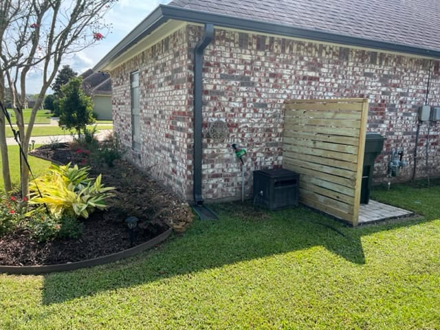 A brick house with a well-maintained flowerbed along its side, featuring green and yellow plants. A wooden privacy screening partially covers garbage bins on a concrete pad. A rain gutter runs down from the roof. The yard is green and neatly trimmed.