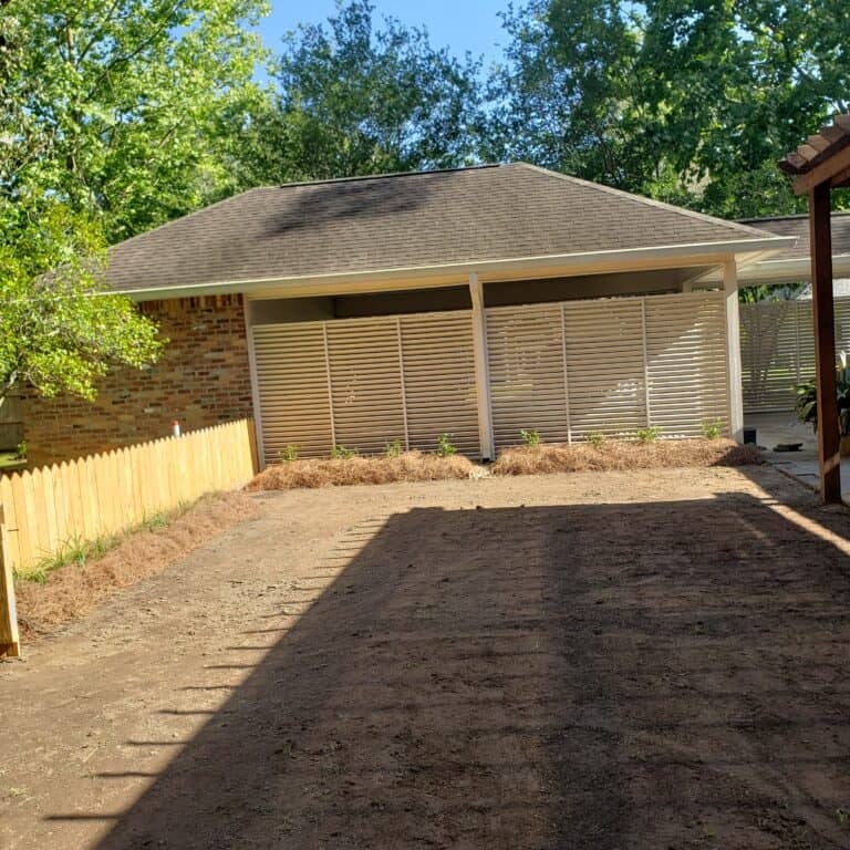 A fenced backyard with freshly tilled soil and a few new plants along the back fence. In the background, there is a brick house with a covered patio and trees providing shade. The area appears prepped for gardening or new lawn installation after some recent gradework.