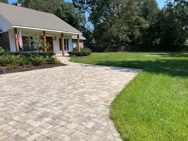 A single-story house with a gray shingle roof, a front porch, and an American flag displayed near the entrance. The house features masonry brick exterior, a paved driveway and walkway leading to the door, and a well-maintained lawn with trees in the background.