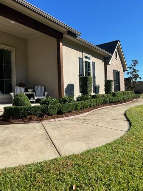 A well-maintained front yard of a house with beige walls and a gray roof. The yard features a curved concrete pathway, manicured bushes, neatly trimmed grass, and two white Adirondack chairs on the porch. Consistent maintenance ensures the clear blue sky provides a bright backdrop.