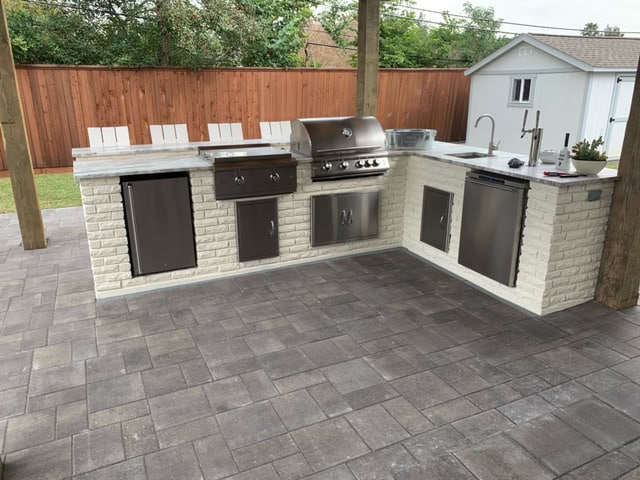 Outdoor kitchen featuring a stainless steel backyard grill, mini fridge, and sink with a white brick base. The patio cooking area is set on grey paver stones and partially covered by a wooden pergola. A wooden fence and small white shed are visible in the background.