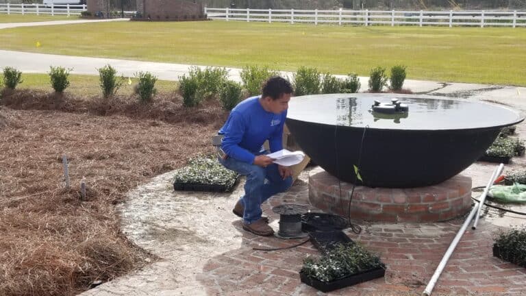 A person in a blue shirt is crouching next to a large, circular water feature on a brick patio. They hold papers and are surrounded by gardening tools and small plants, engaged in garden design. In the background are trimmed hedges, a grassy lawn, and white fences.