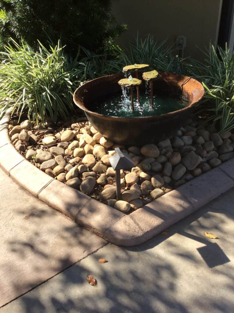 A small round water fountain with three spouts flowing into a rust-colored basin, surrounded by smooth rocks and green plants. This elegant water feature is set in a landscaped garden area adjacent to a paved walkway.
