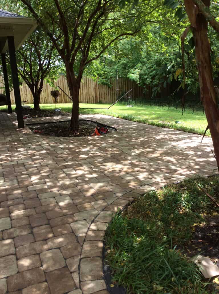 A backyard featuring a neatly paved stone patio area surrounded by trees with dense foliage. A lawnmower and traffic cone are placed on the patio, with a grassy yard and wooden fence in the background. Sunlight filters through the leaves, creating dappled shadows.