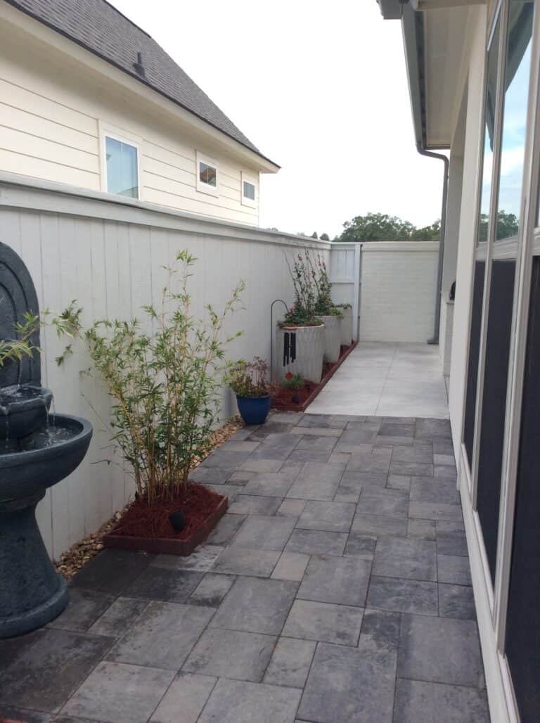 A narrow backyard with a stone tile patio features a wall fountain on the left, two potted bamboo plants, and a plant bed with greenery along the fence. The area is enclosed by a tall wooden fence and adjacent to the side of both a house and a white building.