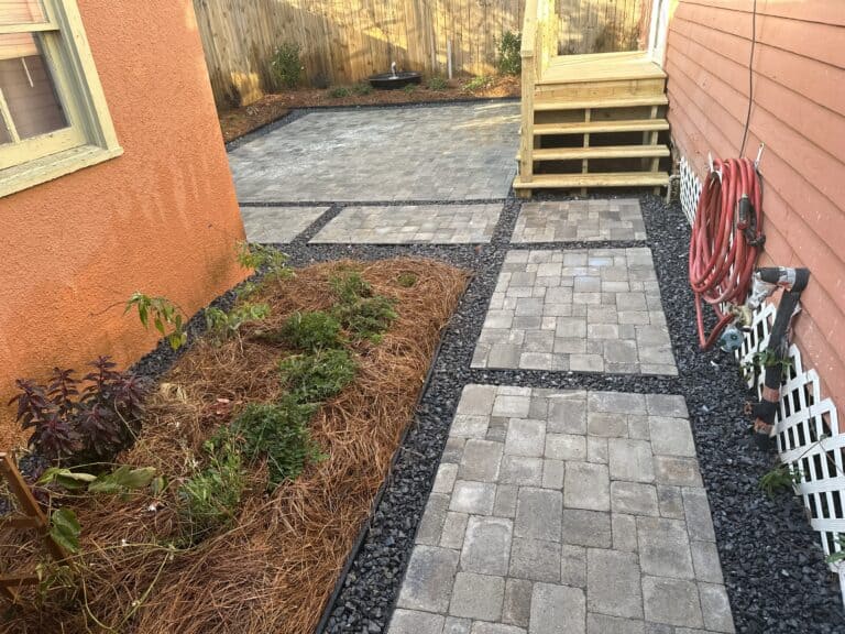 A neatly landscaped backyard features a stone pathway with dark gravel accents, leading to a small patio deck with steps. A freshly mulched garden bed with plants and a coiled red hose attached to the house add to the tidy appearance.