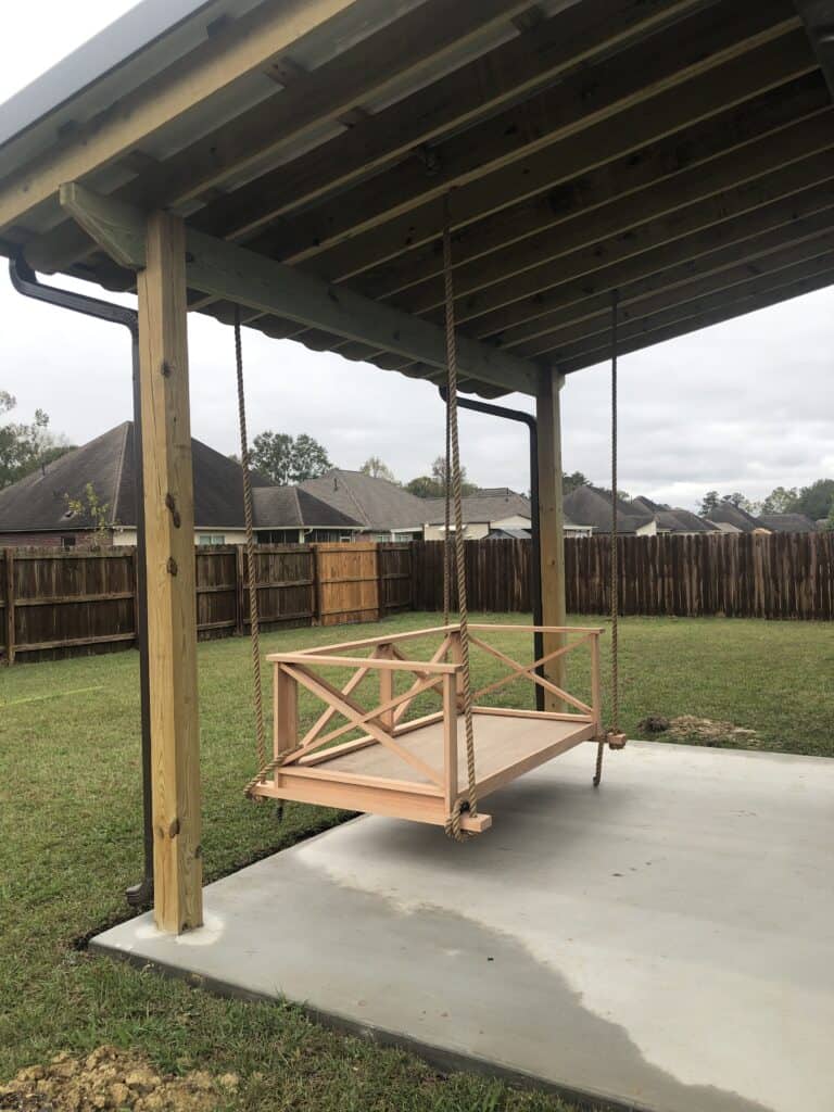 A wooden daybed swing with a flat seat and backrest, suspended by ropes from a covered patio structure. The swing is set on a concrete slab, with a grassy yard and a wooden fence in the background. Several houses are visible beyond the fence under an overcast sky.

