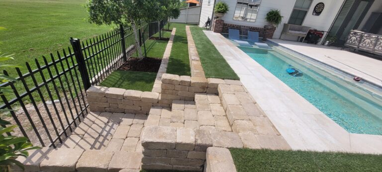 A landscaped backyard featuring a swimming pool with a brick-paved deck. The pool is adjacent to a raised area with synthetic turf supported by stone retaining walls. A black metal fence separates the yard from a grassy field, and two chairs are situated by the pool.