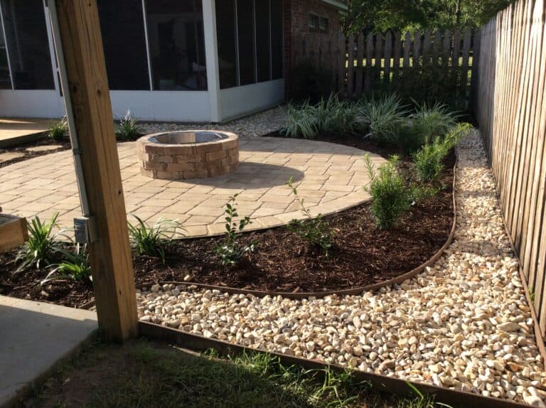 A landscaped backyard displays a fabulous circular stone fire pit on a paved patio area. Surrounding the patio are mulched garden beds with green plants and a border of light-colored rocks. A wooden fence and screened-in porch complete this inviting outdoor space.