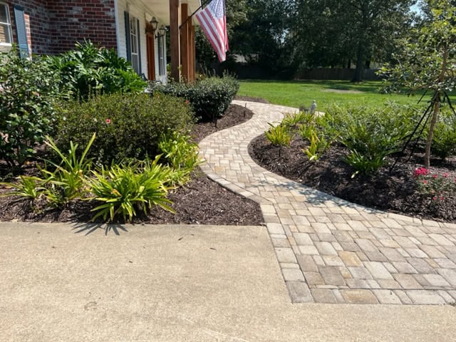 A neatly paved, winding stone walkway bordered by mulch and various plants leads to the front door of a brick house. An American flag is mounted near the entrance, and a lush green lawn surrounds the masonry path and driveway.