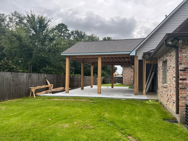 A backyard with a wooden pergola under construction attached to a brick house. The pergola, part of recent roof additions, has a gravel-covered floor and wooden beams supporting a partially finished roof. The fenced yard boasts green grass, and construction materials are placed on the side.