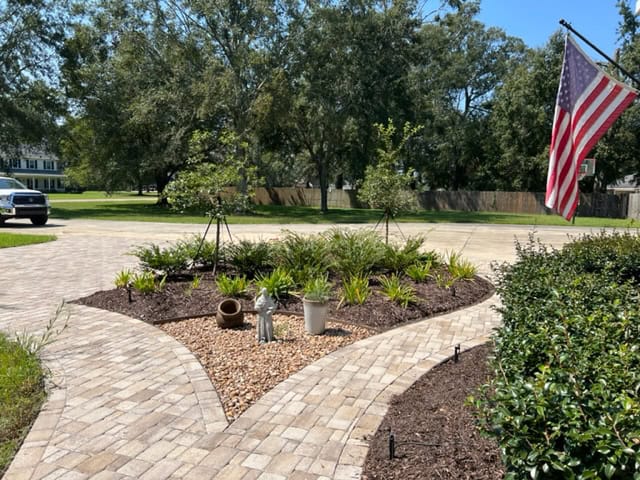 A front yard garden with a circular stone pathway, featuring neatly arranged plants, a small statue, and garden decor. An American flag is mounted on a pole to the right, and a white SUV is visible in the background near the masonry driveway by the street.