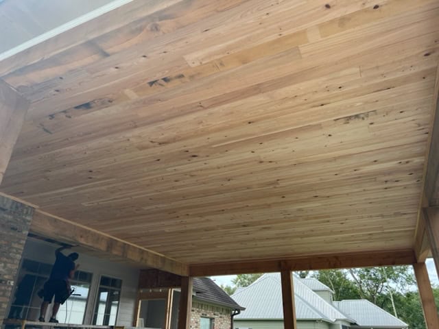 A worker stands on a scaffold, installing or inspecting roof extensions for a wooden ceiling in an outdoor patio area. The ceiling consists of neatly aligned wooden planks. In the background, you can see partial views of houses and trees under a cloudy sky.