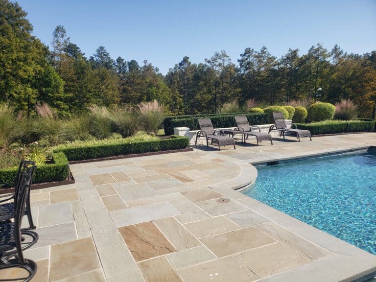 A serene outdoor pool area with stone paving, surrounded by lounge chairs and manicured greenery, showcases impeccable maintenance. The pool water is a clear blue, and lush trees are visible in the background under a clear sky.