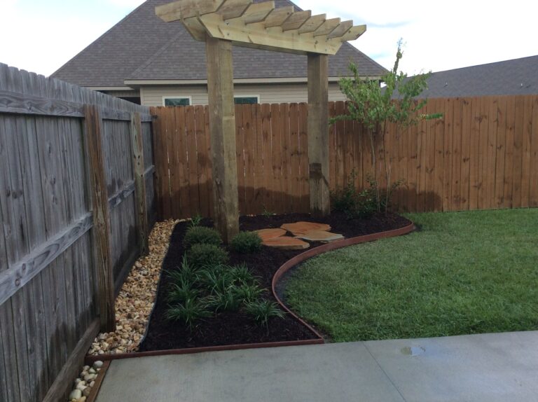 A backyard garden featuring a wooden pergola, various arbors, a mulched flowerbed with shrubs and plants, a stone pathway, and a strip of decorative rocks along the wooden fence. The garden is bordered by a neatly mowed lawn and a concrete patio, showcasing elegant outdoor decor.