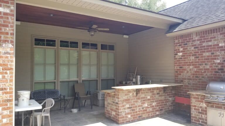 A partially shaded patio with a ceiling fan, various chairs, a table, and a brick masonry outdoor kitchen area. The outdoor kitchen includes a grill, sink, and counter space. The brickwork on the patio matches the house exterior perfectly.