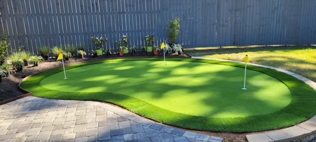A backyard scene featuring one of the finest putting greens with three holes, each marked by a yellow flag. The putting green is surrounded by a stone paver path, with various plants and shrubs lining the wooden fence in the background.