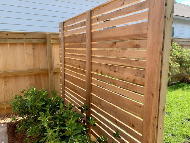 A wooden privacy screen stands in a backyard, featuring horizontal slats and positioned near a corner with a fence. Green shrubs grow in front of the screening, and the background includes a neighboring house and a grassy lawn.