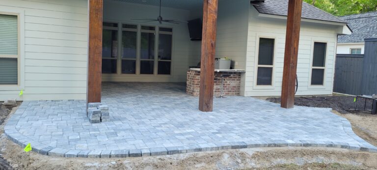A covered outdoor patio with gray stone pavers and wooden support beams. An outdoor kitchen with a brick base includes a grill and a countertop. The patio, nestled in the backyard of a beige house with large windows and a door, overlooks an undeveloped yard area.