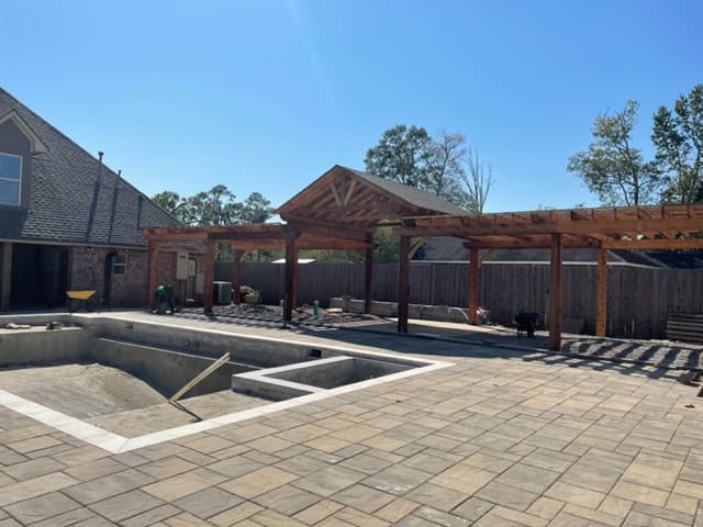 A backyard scene featuring a partially constructed in-ground pool, surrounded by a large stone patio. Adjacent to the pool are wooden pergolas under construction. A house and a wooden fence are visible in the background under a clear blue sky.