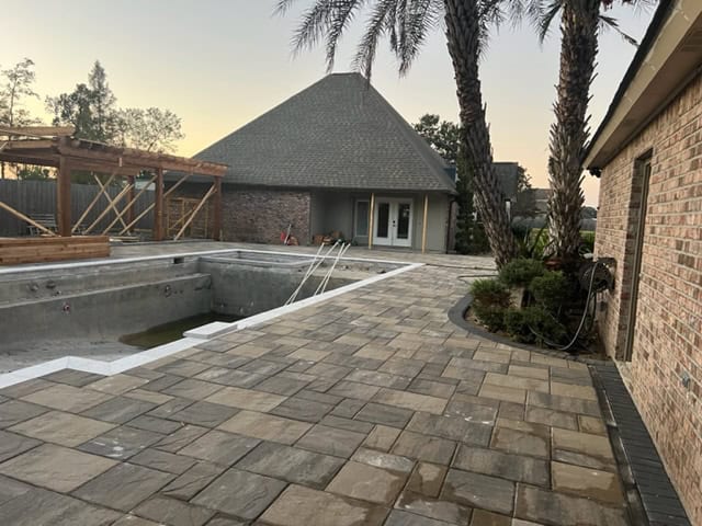 A large backyard with an empty in-ground swimming pool under construction features palm trees and a stone patio area. A small, roofed structure with a triangular shape is in the background, near the pool.