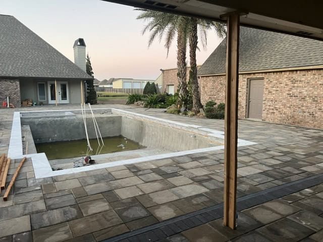 A backyard under renovation features an empty rectangular pool surrounded by a freshly paved stone patio. The area includes palm trees and modern brick buildings. Construction materials are visible near the pool, indicating ongoing work on the patio.